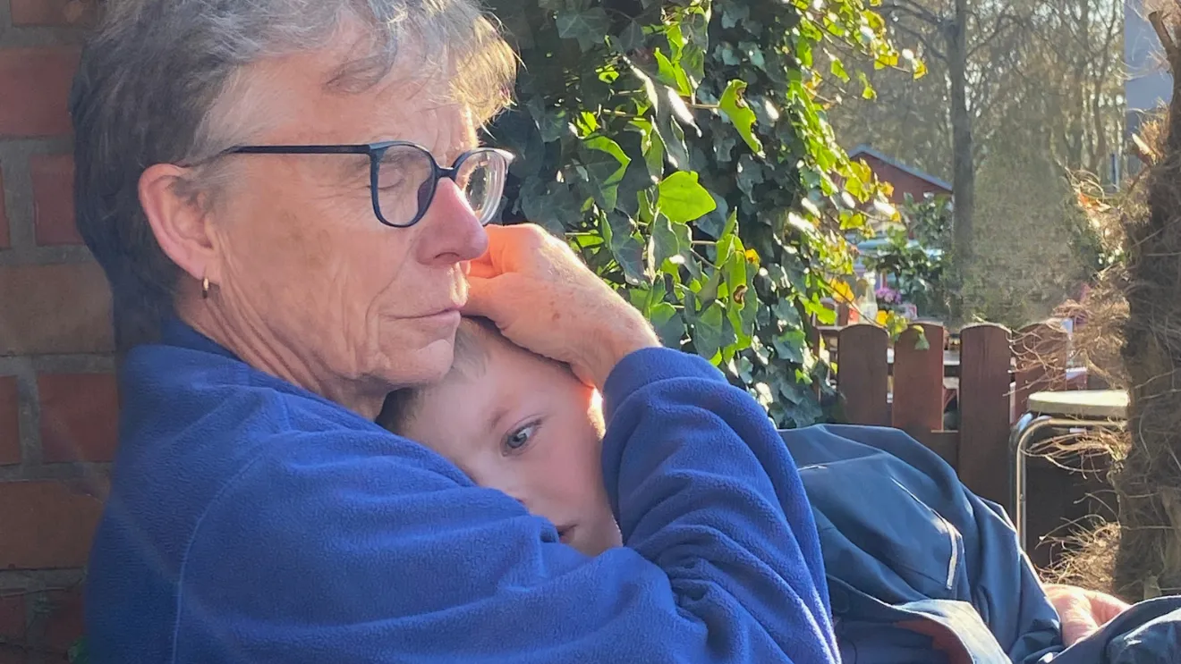 Eine ältere Person mit Brille umarmt ein Kind herzlich; sie sitzen im Freien im Sonnenlicht, nahe einer Backsteinmauer und grünem Blattwerk.