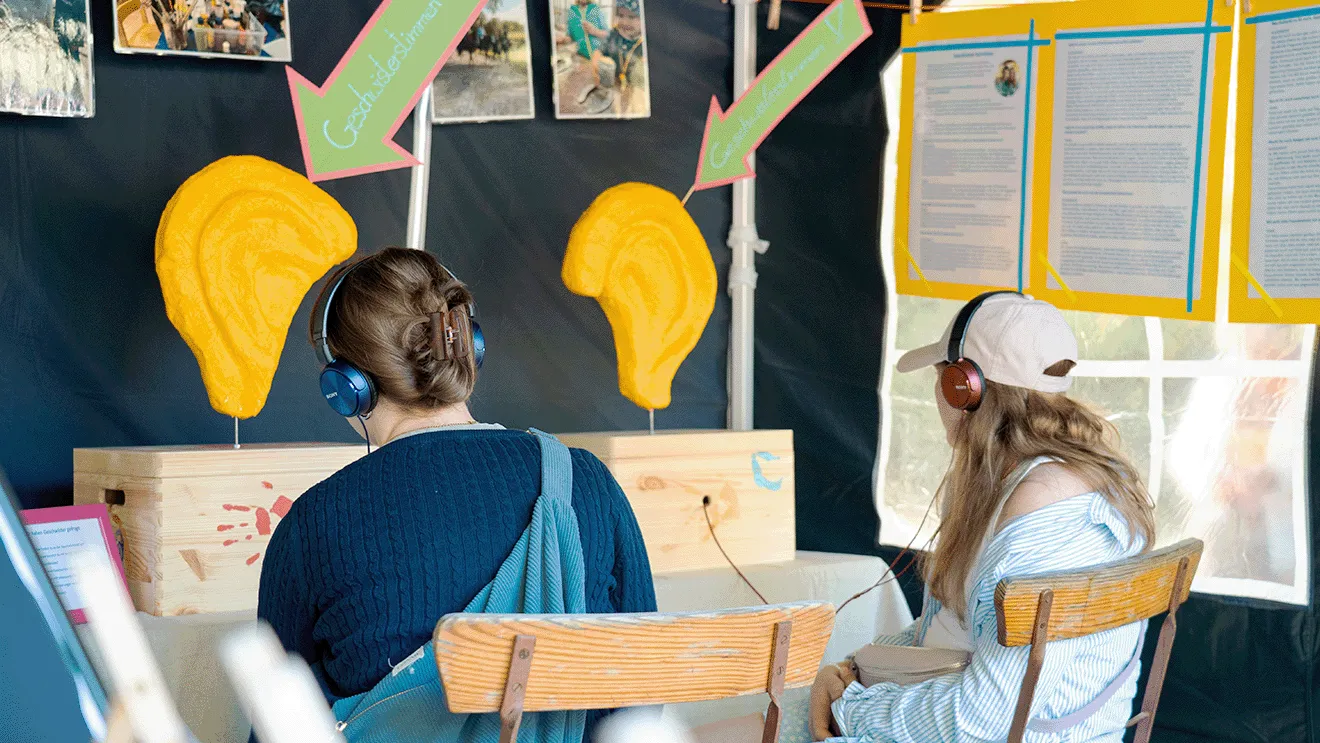 Zwei Personen mit Kopfhörern sitzen vor einem Schaukasten mit großen gelben Ohrmodellen, umgeben von Informationstafeln und Fotos.