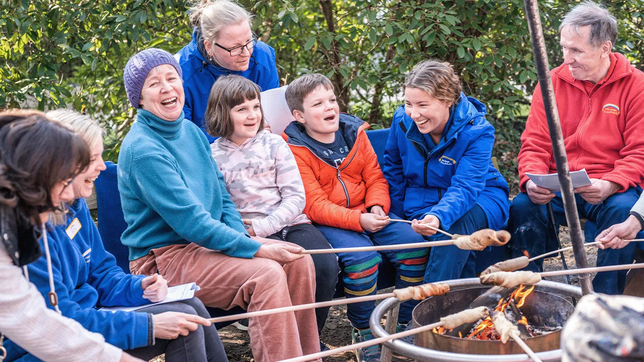 Eine Gruppe von Menschen, darunter auch Kinder, sitzt um ein Lagerfeuer herum, lächelt und röstet im Freien Essen auf Stöcken.