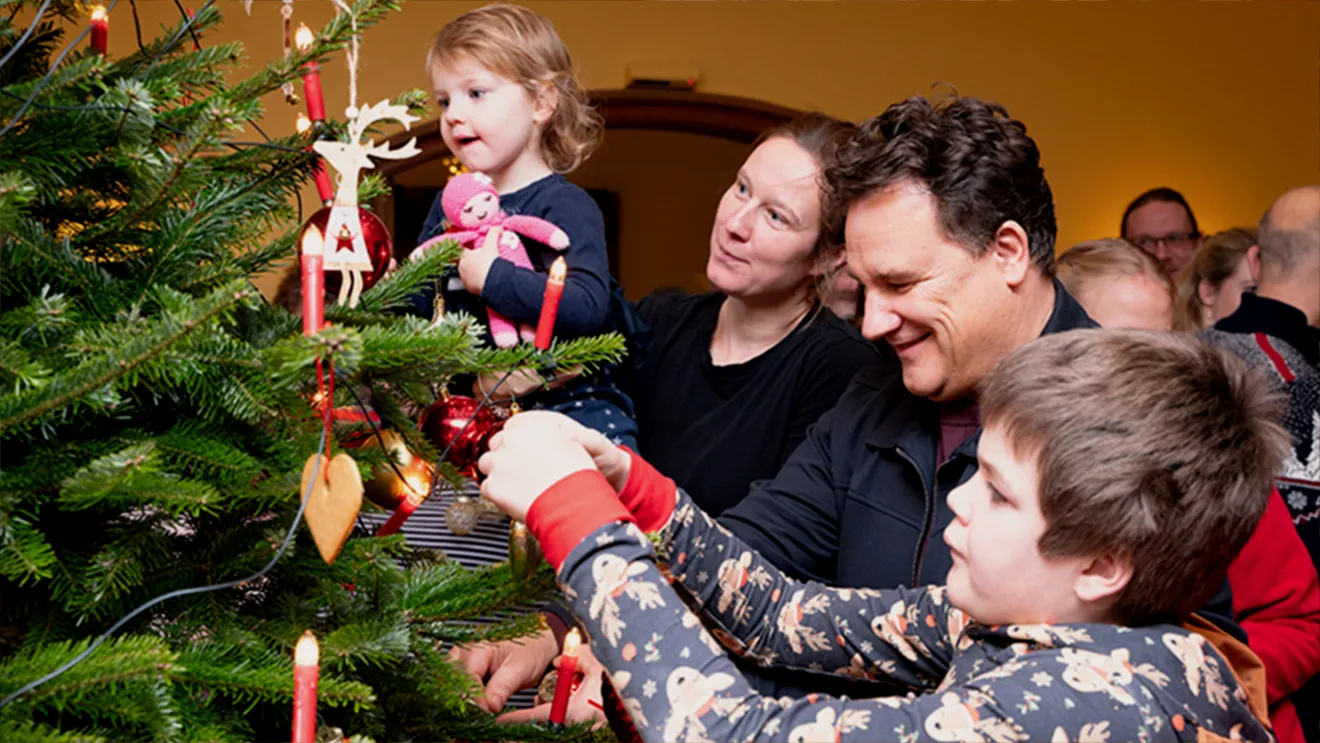 Familie schmückt einen Weihnachtsbaum mit Ornamenten und Kerzen; ein Kind wird hochgehoben, um eine Dekoration anzubringen.