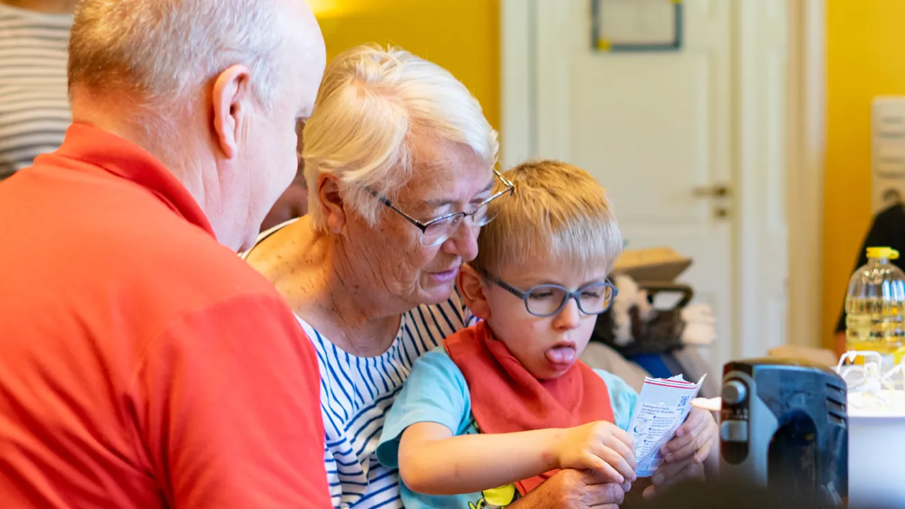 Ältere Menschen und ein kleiner Junge lesen gemeinsam eine Broschüre an einem Tisch in einem hell erleuchteten Zimmer. Der Junge wirkt konzentriert und trägt eine Brille.