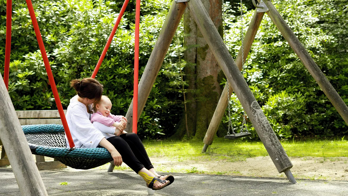 Frau sitzt auf einer Schaukel mit einem Baby auf dem Schoß, umgeben von Grünpflanzen in einer Parklandschaft.