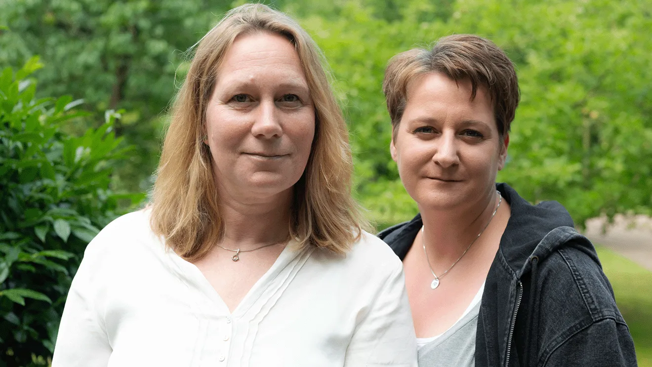 Zwei Frauen stehen im Freien, im Hintergrund ist Grün zu sehen. Die eine trägt ein weißes Hemd, die andere ein graues Oberteil mit einer dunklen Jacke.