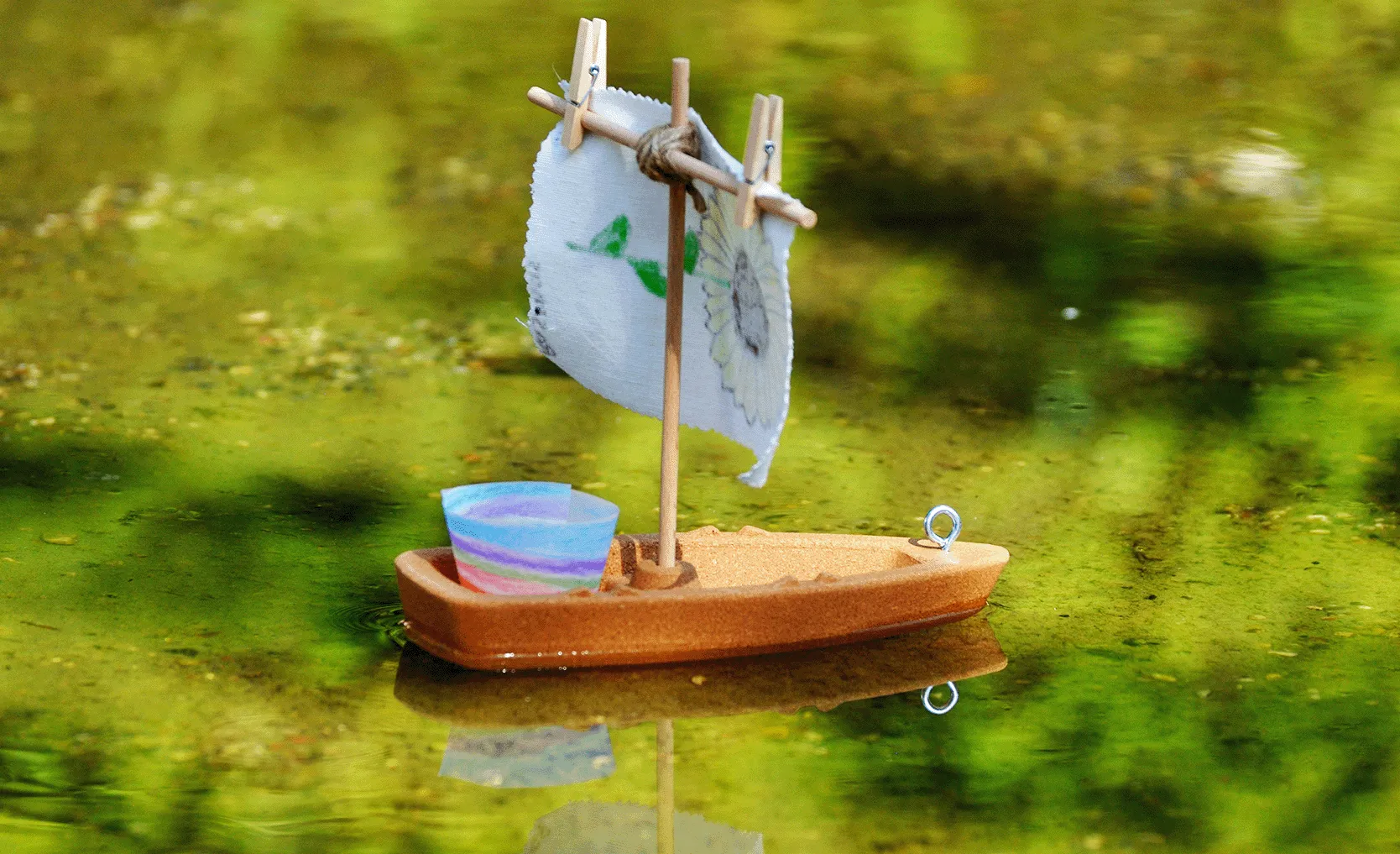 Ein kleines Spielzeugsegelboot mit einem bunten Becher und einem Stoffsegel schwimmt auf einer spiegelnden grünen Wasseroberfläche.
