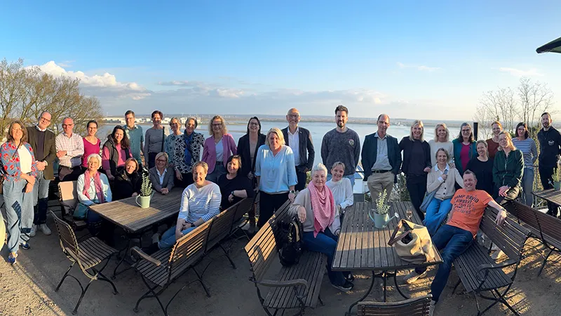 Eine Gruppe von Menschen versammelte sich im Freien und posierte für ein Foto auf einer Terrasse mit Holztischen und -stühlen unter einem strahlend blauen Himmel.
