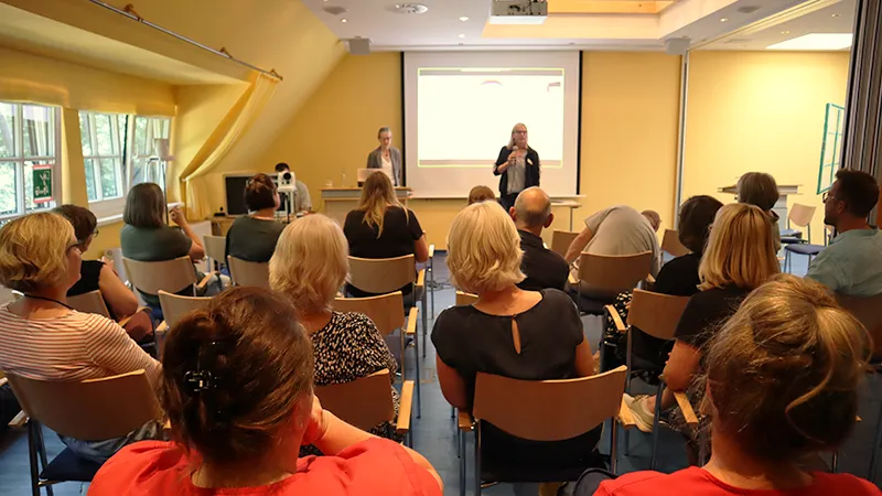 Eine Gruppe von Personen sitzt in einem Raum und blickt auf zwei Referenten, die vor einer Leinwand stehen und eine Präsentation halten.