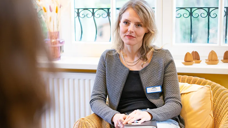 Psychologische Begleitung eine blonde Frau sitzt im Sessel hört einer anderen Person, die man verschwommen im Vordergrund sieht, zu.