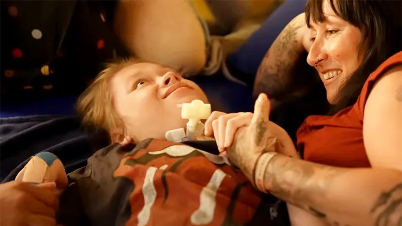 Eine lächelnde Frau mit Tätowierungen hält die Hand eines liegenden Kindes, das zu ihr aufschaut. Das Kind trägt ein T-Shirt mit einem grafischen Aufdruck.