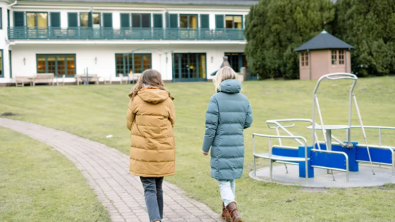 Zwei Personen in Wintermänteln gehen einen Weg in einem Park entlang. Im Hintergrund sind ein Spielplatz und ein großes Gebäude zu sehen.