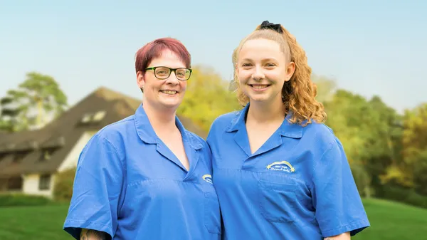 Two smiling people in blue uniforms are standing outdoors, with a blurred background of a house and trees under a clear sky.