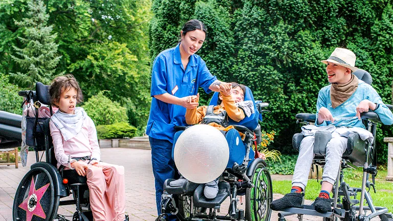 Eine Pflegekraft interagiert mit drei Kindern im Rollstuhl im Freien und beschäftigt sie mit einem großen Ballon. Im Hintergrund sind Bäume und Grünflächen zu sehen.