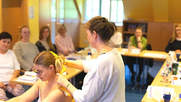 Mehrere Personen sitzen an Tischen in einem Seminarraum. Eine Frau steht und legt einer sitzenden Frau ein gelbes Handtuch um den Nacken.