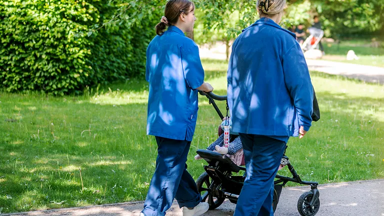 Two adults in blue work clothing are pushing a stroller through a garden. A child is lying in the stroller.