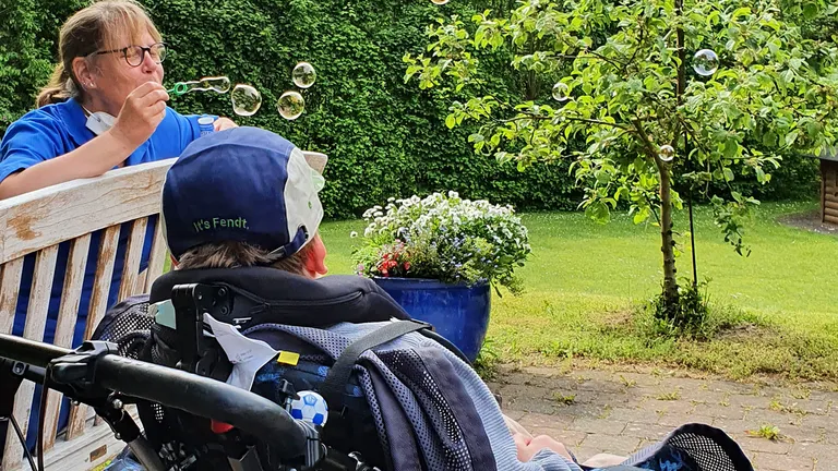 Eine Pflegekraft bläst Seifenblasen in der Nähe einer Person im Rollstuhl in einem Garten, mit einem Baum und Blumen im Hintergrund.