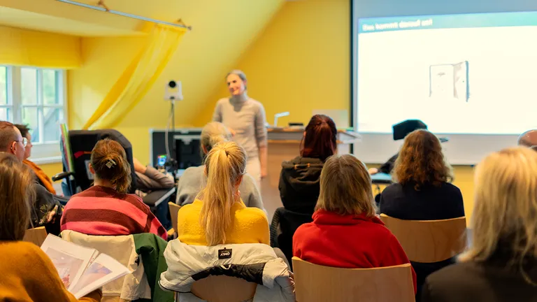 A woman gives a presentation to an audience in a bright room with yellow walls. A projector screen displays content in the background.