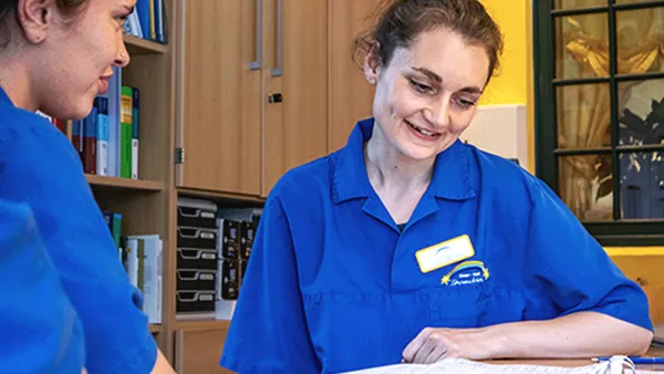 Zwei Mitarbeiter des Gesundheitswesens in blauen Uniformen besprechen Papierkram an einem Schreibtisch in einem hell erleuchteten Büro mit Regalen und einem Fenster im Hintergrund.