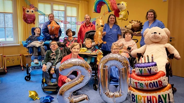 Group of people, including children in wheelchairs and adults, with colorful balloons and a large "20" balloon, celebrating a birthday.