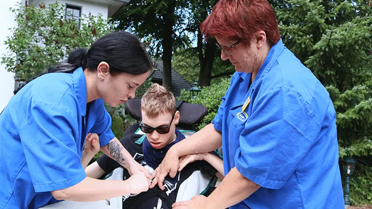 Zwei Pflegekräfte in blauen Uniformen helfen einem jungen Menschen im Rollstuhl im Freien, umgeben von Bäumen und einem Gebäude.