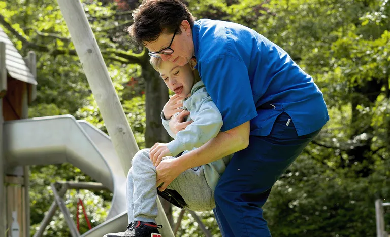 Eine Betreuerin in blauer Kleidung hilft einem Kind auf einer Schaukel in einem sonnigen Spielplatz mit einer Rutsche und Bäumen im Hintergrund.