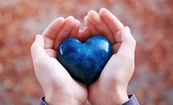 Hände, die sanft einen polierten blauen herzförmigen Stein vor einem verschwommenen, warmtonigen Hintergrund halten.