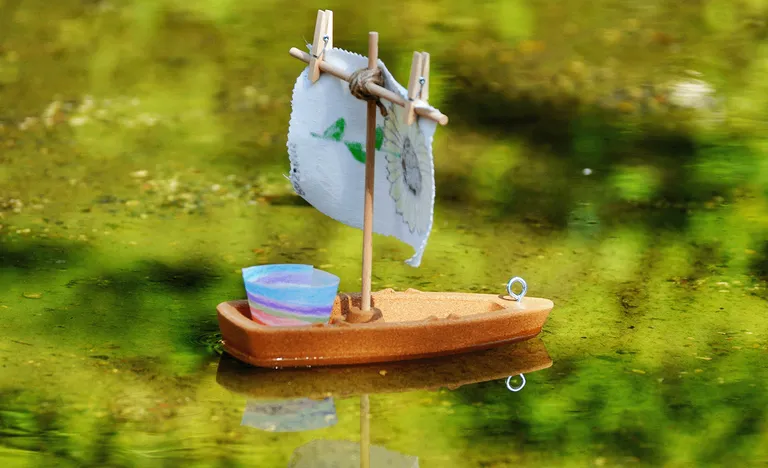 Ein kleines Spielzeugsegelboot mit einem bunten Becher und einem Stoffsegel schwimmt auf einer spiegelnden grünen Wasseroberfläche.