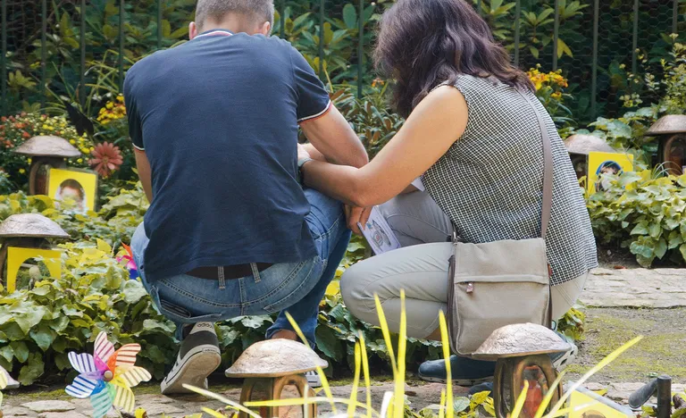 Zwei Menschen knien in einem Garten, umgeben von Pflanzen und kleinen Dekorationselementen, darunter bunte Windräder und gerahmte Fotos.