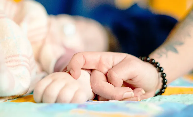 Eine erwachsene Hand berührt die Hand eines Kindes. Beide liegen auf einer bunten Decke. Der Arm der erwachsenen Person ist tätowiert und trägt ein schwarzes Perlenarmband.