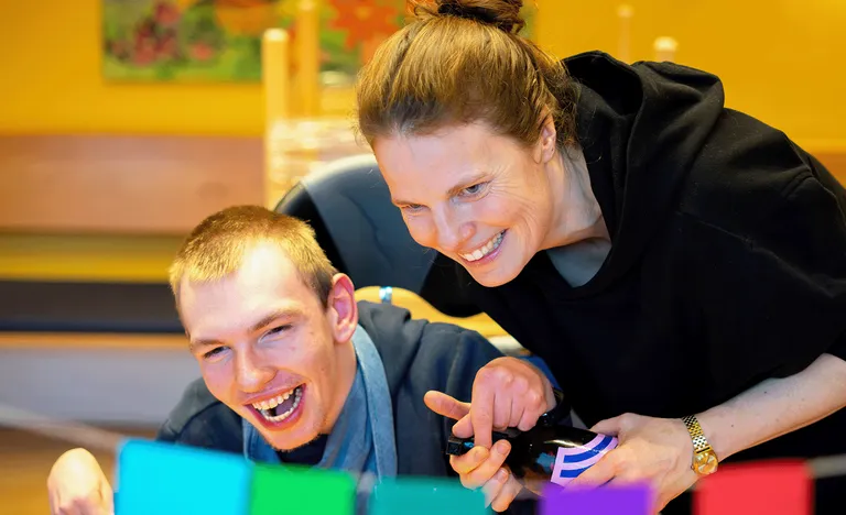 Eine lächelnde Frau und ein Mann sind in einem Innenraum mit einer farbenfrohen Aktivität beschäftigt. Der Hintergrund ist in warmen Farbtönen gehalten, mit unscharfen Details.