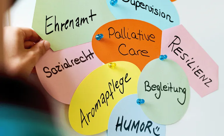 Bunte Papierformen mit handgeschriebenen deutschen Wörtern wie „Ehrenamt“, „Palliative Care“ und „Resilienz“, die an eine Tafel geheftet sind.