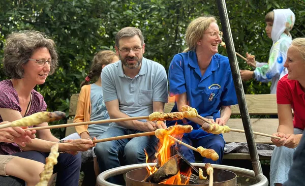 Eine Gruppe von Menschen sitzt um ein Lagerfeuer herum, lächelt und röstet Brot auf Stöcken, im Hintergrund ist Grün zu sehen.