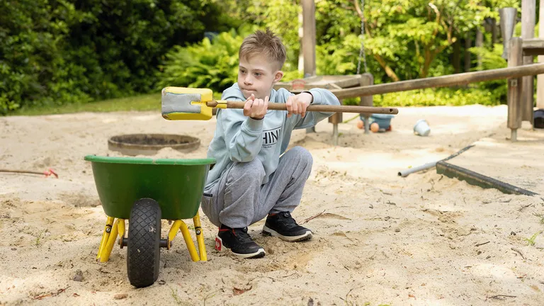 Ein Junge sitzt in einem Sandkasten und hält einen gelben Spielzeugspaten mit langem Stiel. Vor ihm steht eine grüne Schubkarre.