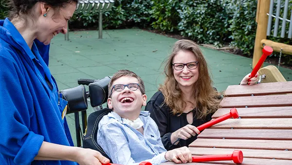 Three people joyfully play a large outdoor xylophone. A child in a wheelchair laughs while two adults smile, surrounded by greenery.