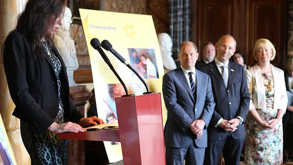 A woman speaks at a podium with a microphone, while four people stand attentively near a display board in an elegant room.