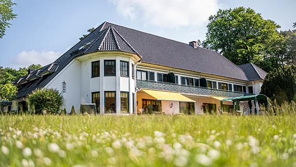 Großes weißes Haus mit dunklem Dach, umgeben von grünen Bäumen und Gras. Einige Fenster sind mit Markisen versehen. Es ist ein heller, sonniger Tag.