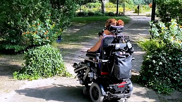 Eine Person in einem motorisierten Rollstuhl fährt an einem sonnigen Tag auf einem Gartenweg entlang, umgeben von Grünpflanzen und Blumen.
