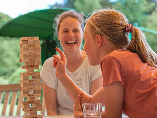 Ein Mädchen entfernt vorsichtig einen Block aus einem Jenga-Turm, während eine Frau im Hintergrund lächelt. Sie befinden sich im Freien unter einem grünen Sonnenschirm.