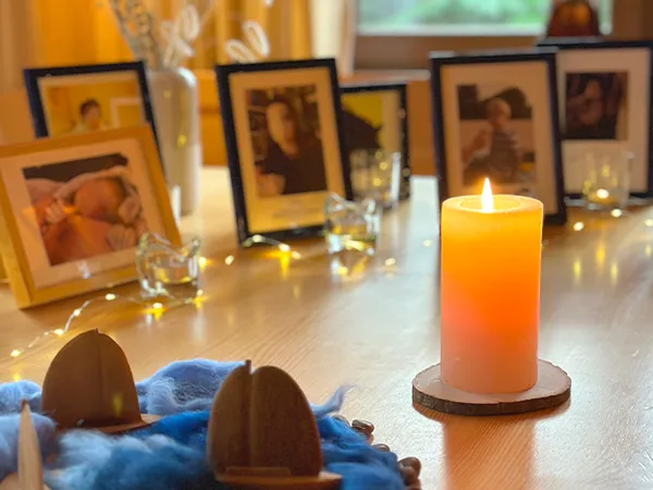 A lit candle on a wooden table surrounded by framed photos, decorative items, and soft lighting, creating a warm, cozy atmosphere.