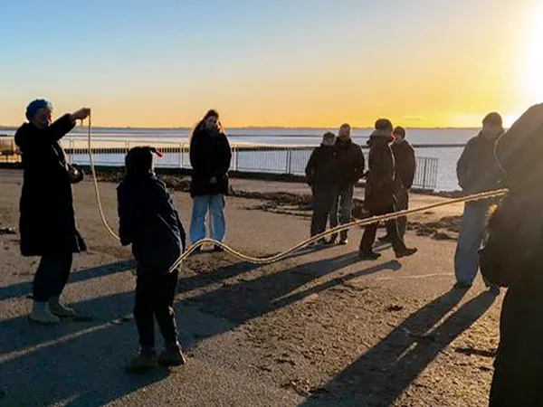 Menschen, die an einem sonnigen Tag am Wasser Tauziehen spielen und lange Schatten auf den Boden werfen.