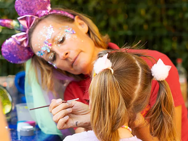 Eine Frau mit glitzernder Gesichtsfarbe und violetten Ohren bemalt das Gesicht eines jungen Mädchens bei einer Veranstaltung im Freien.