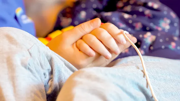 Two hands gently holding each other, one with an IV line attached. The background is softly blurred, featuring a patterned fabric.