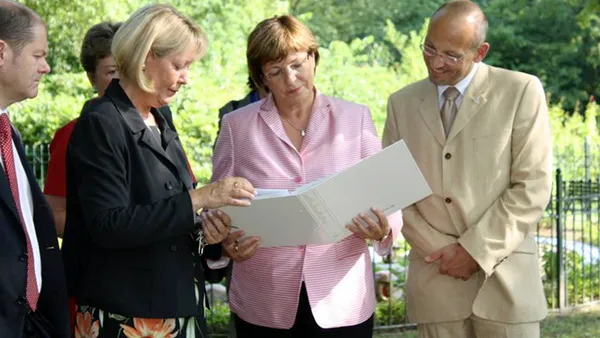 A group of people outdoors examining a document together, with greenery in the background.
