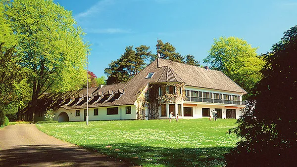 A large, traditional house with a sloped roof surrounded by lush greenery and trees under a clear blue sky. People are walking on the lawn.