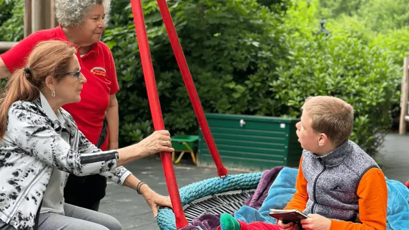 A child sits on a swing holding a tablet, while two adults, one standing and one kneeling, engage with him in a park setting.