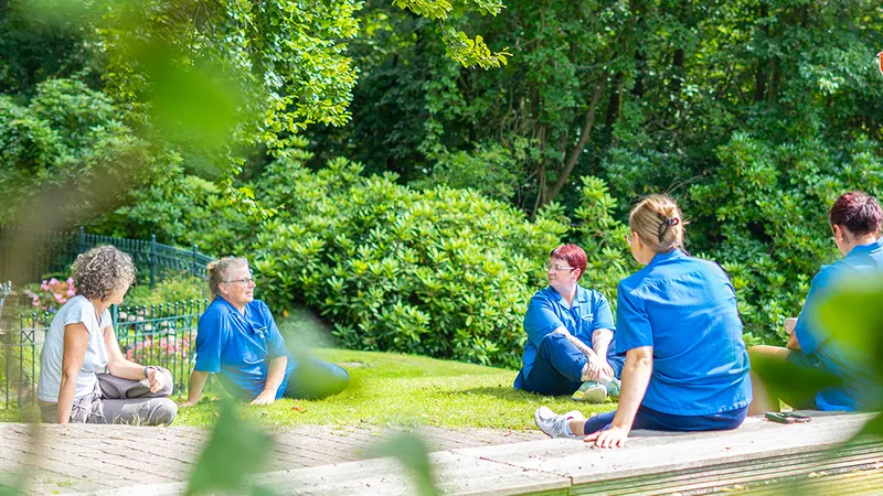 Eine Gruppe von Menschen in blauen Hemden sitzt auf dem Rasen in einem sonnigen Garten, umgeben von Bäumen und Grünpflanzen, und unterhält sich.
