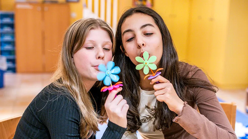 Two girls blowing on colorful pinwheel toys indoors, smiling and enjoying the moment.