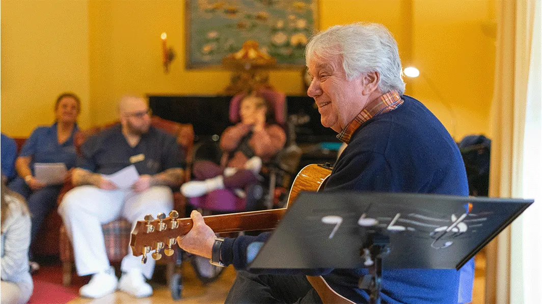 Rolf Zuckowski lächelt und spielt Akustikgitarre für ein sitzendes Publikum in einem gemütlichen Raum, Notenblatt auf einem Ständer im Vordergrund.