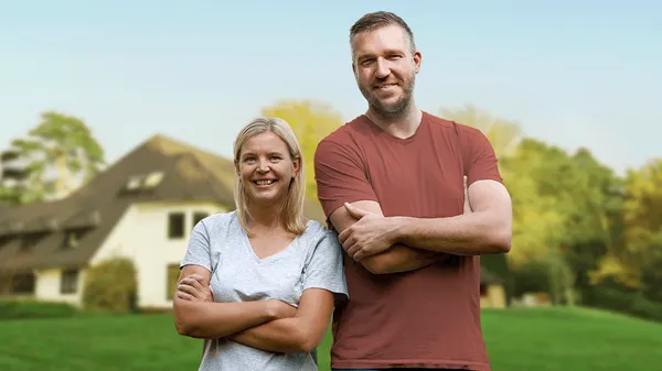 Ein Mann und eine Frau stehen lächelnd mit verschränkten Armen auf einer grünen Wiese vor einem Haus im Hintergrund.