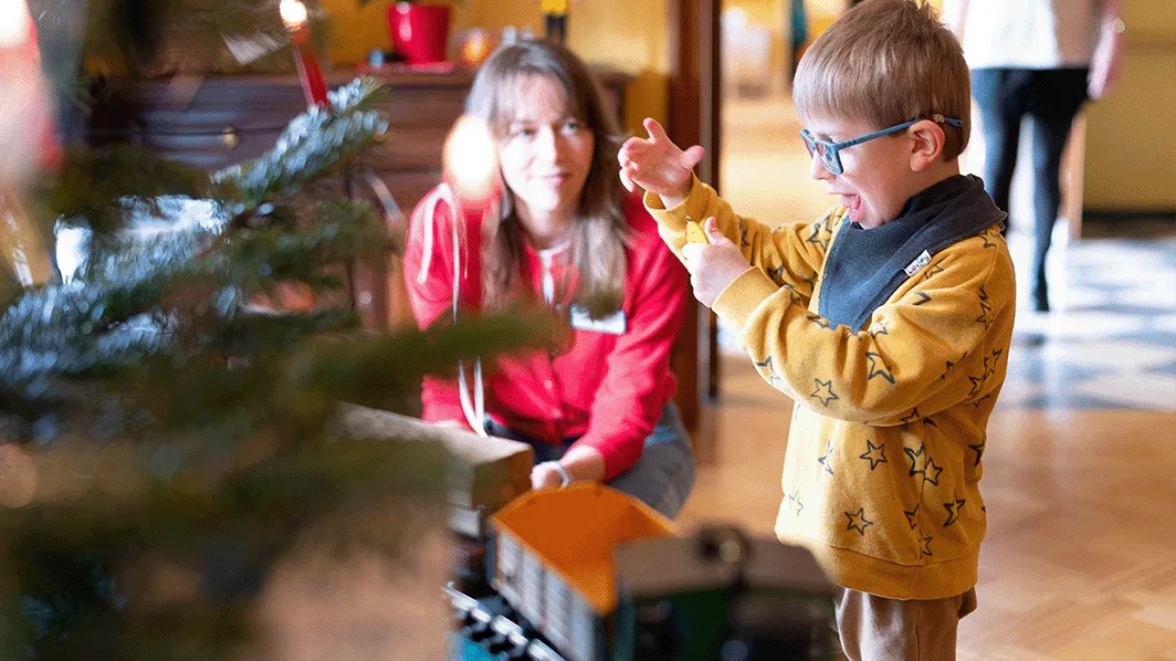 Ein Kind mit Brille spielt begeistert mit einer Spielzeugeisenbahn, während eine Frau in einem roten Pullover in einem Zimmer mit einem Weihnachtsbaum zusieht.