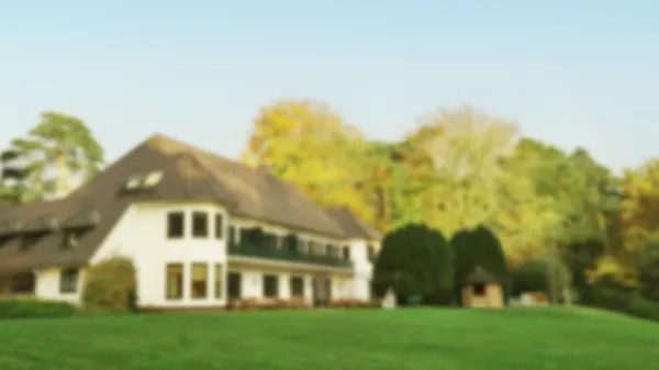 Unscharfes Bild eines großen Hauses mit braunem Dach, umgeben von einem grünen Rasen und buntem Herbstlaub.