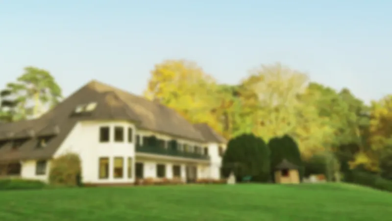 Unscharfes Bild eines großen Hauses mit braunem Dach, umgeben von einem grünen Rasen und buntem Herbstlaub.